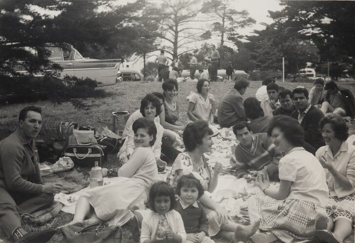 Large group of men, women and children picnicking in a park. Car and trees in background.