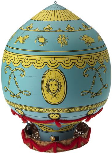 Blue wooden balloon model with yellow patterns. Basket base holds figurines. Side A.