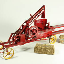 Red metal hay press model with gears, springs, four yellow wheels, white painted letters. Two hay bales nearby