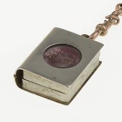 Detail of silver watch chain with miniature book in silver case with cut-out circle. Closed.