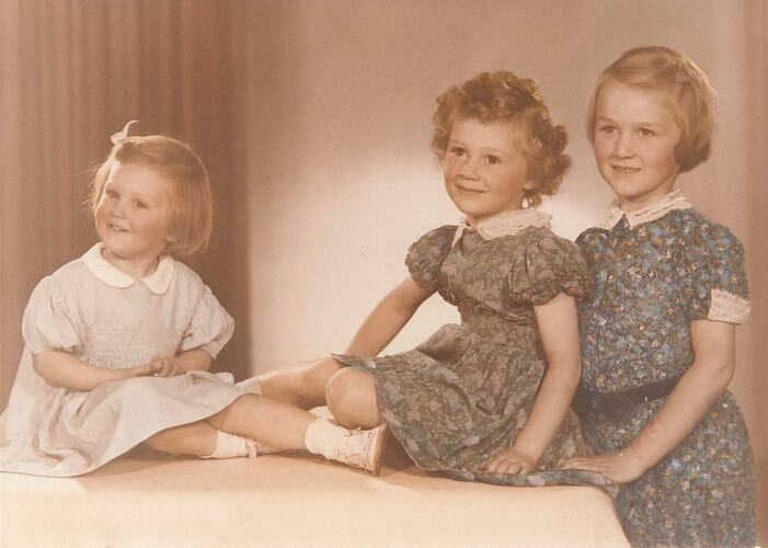 Two young girls sitting on a bench. A third older girl stands beside them.