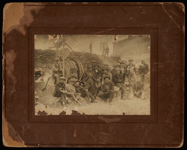 Group of construction workers pose on rocky worksite with machinery behind them.