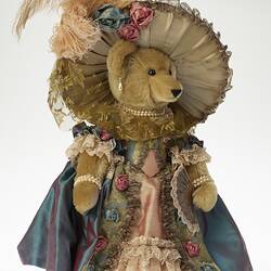 Light brown plush bear wearing lavish cream hat with feather and ornate dusty pink dress and blue jacket.