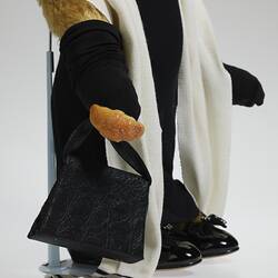 Detail of light brown bear. Wears black dress, full-length gloves, handbag and shoes, white pearls and scarf.