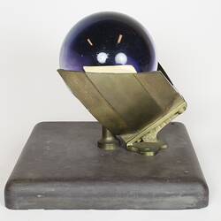 Purple glass sphere sitting on metal frame with square base, side view.