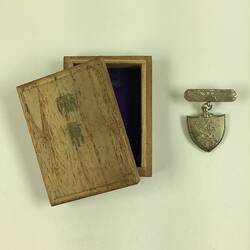 Wooden lid with two black stamped characters on base of wooden box. Shield medal beside it.