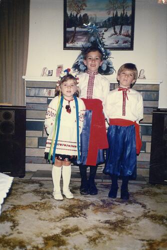 Three children dressed in Ukrainian national dress pose in front of a slate mantel with heater.