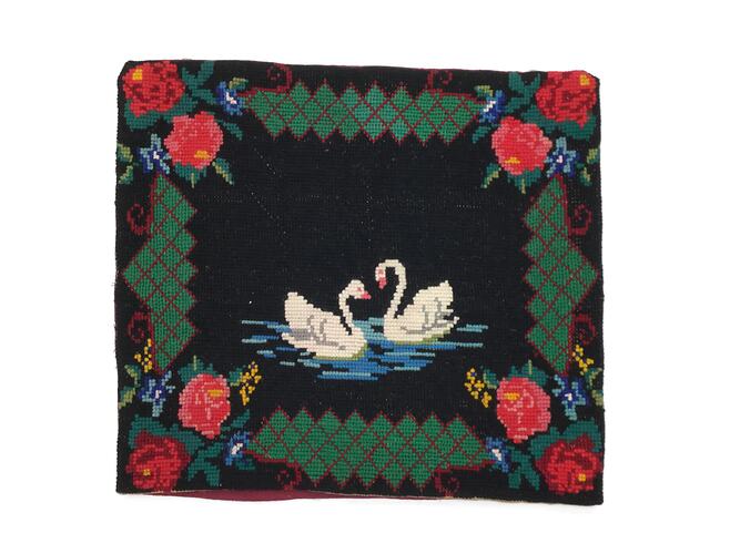 Cushion cover embroidered with two white swans on blue water within a border comprising two red roses and other floral motifs in each corner connected by a margin of diamond shapes in grey-green thread outlined in red, all on a black background.