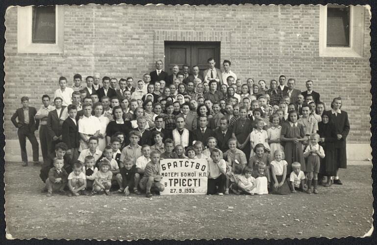 Large group (over 50) pose in front of brick building. Front centre seated children hold a sign.