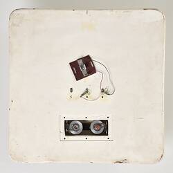 Scuffed white plastic square plate with curved base and rounded corners. Small opening on face side up has two round metal discs within. Above it some wires and a small metal plate are attached to surface.