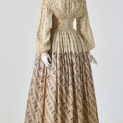 Cream muslin dress, high neck, long sleeves, banded waist, full length patterned, gathered skirt.