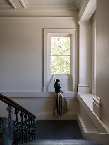 Woman in dark top and pale long skirt climbing a wall up to a window in a staircase.
