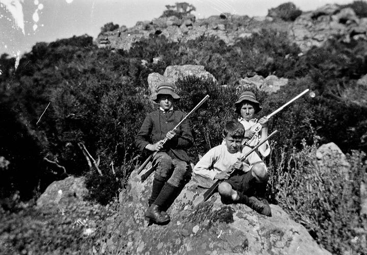 Three young boys with rifles seated on a rocky outcrop.
