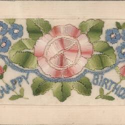Card with embroidered text and pink flower with green leaves in centre, small blue flowers surrounding.