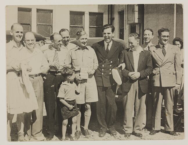 Nine men in suits and one boy, standing in front of a building.
