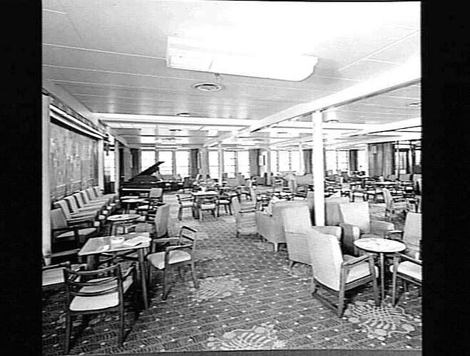 Ship interior. Room with upholstered chairs and armchairs around tables. Piano in background. Lounge area.