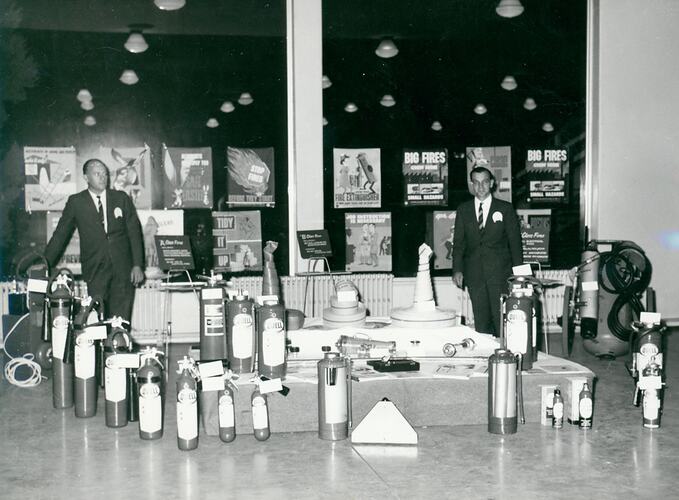 Two men stand at the back of a display of fire fighting equipment.