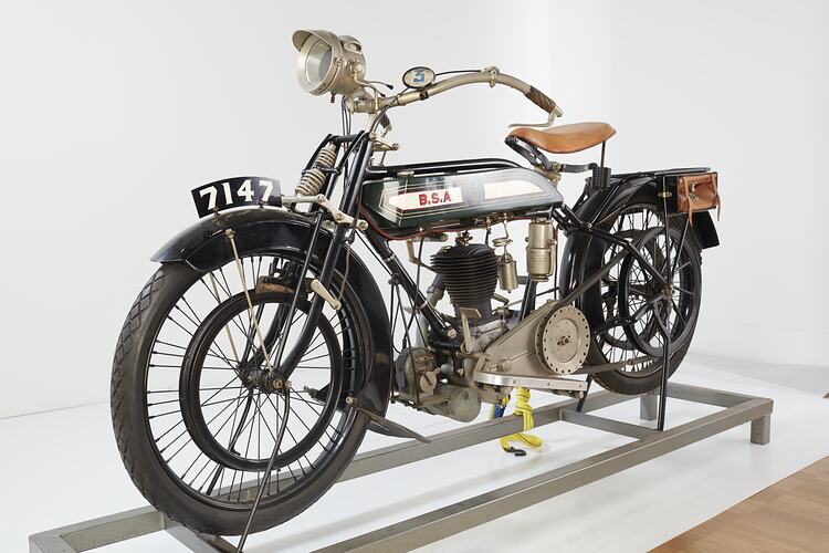 Green, cream motor cycle. Black fenders. Single lamp, brown leather seat, satchel on back.