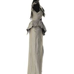 Evening dress, full length, silver-grey silk. No sleeves, winged shoulder peaks, ruffle at waist. Profile.