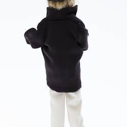 Miniature man in dark coat, light pants. Has a hat and fair hair. Back view.