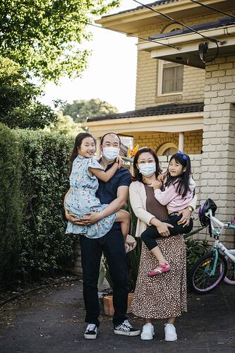 Woman, man and two girls in front of brick house. The woman and man wear masks and each holds a child.