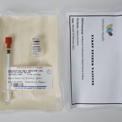 Labelled syringe and vial in lined plastic box. Card in lid has printed text. White background.