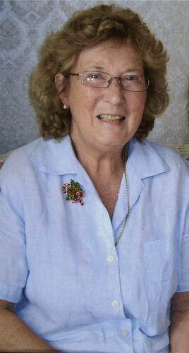 Woman smiling. She wears a blue blouse, glasses and christmas themed brooch.
