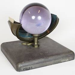 Purple glass sphere sitting on metal frame with square base, three quarter view.