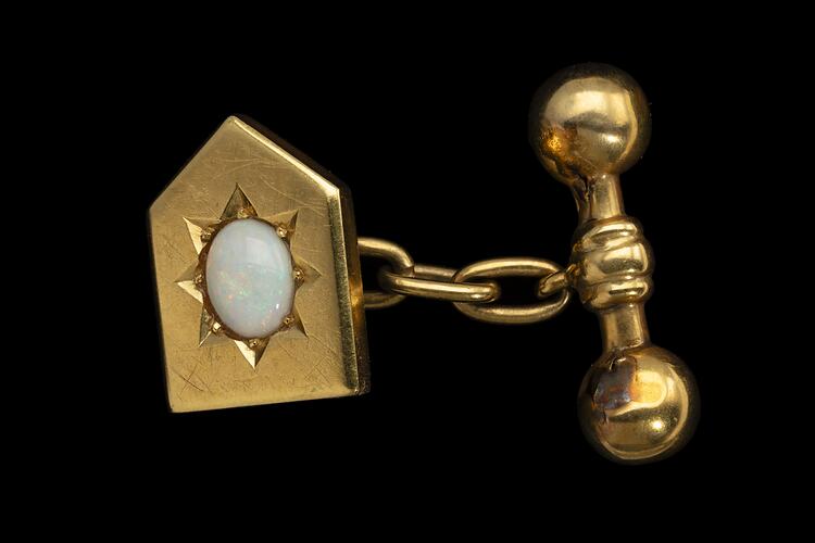 Gold cufflink of heraldic shield design. Centrally placed opal. At back, a chain links it to a barbell.