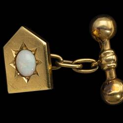 Gold cufflink of heraldic shield design. Centrally placed opal. At back, a chain links it to a barbell.