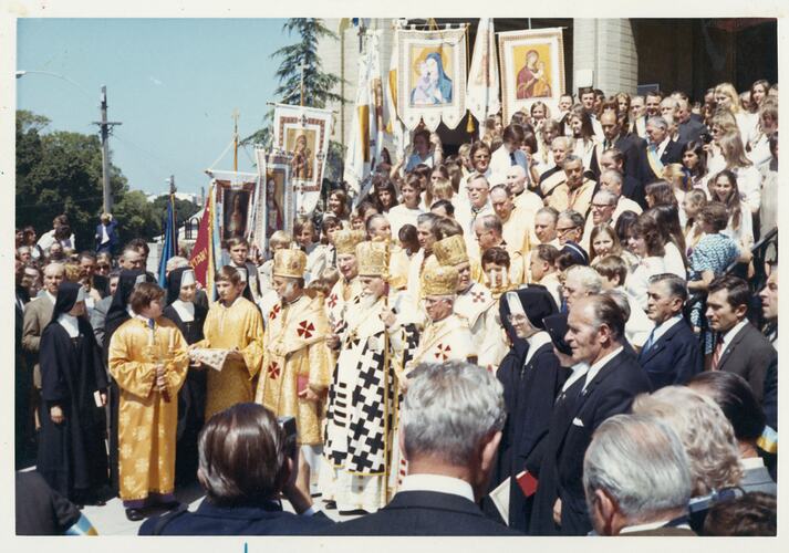 Church congregation welcoming Ukrainian Cardinal and group in gold yellow regalia. Crowds fill church steps. Flags in background.