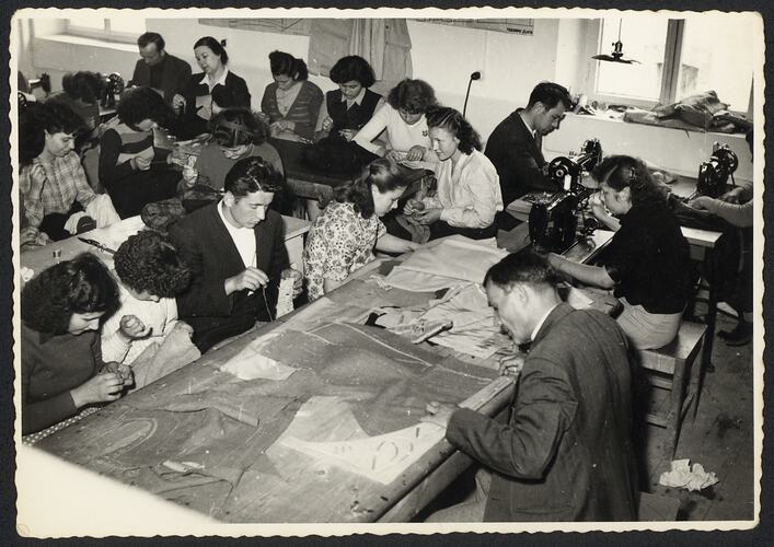 Male and female refugees sit at tables working in a tailoring workshop.