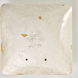 Scuffed white plastic square plate with curved base and rounded corners. Small opening on face side up has two round metal discs within. Above it some wires and a small metal plate are attached to surface.