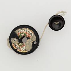 Black wooden cotton reel with two nails in top. White string tied around the middle. The string has a black button on the end. Top view.