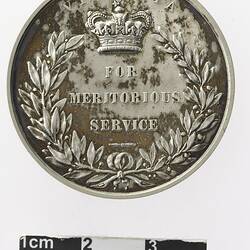 Round silver coloured medal with crown above text and wreath surrounding.