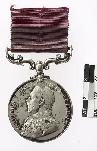 Silver coloured medal with profile of a man and text surrounding, with maroon ribbon.