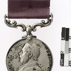 Silver coloured medal with profile of a man and text surrounding, with maroon ribbon.