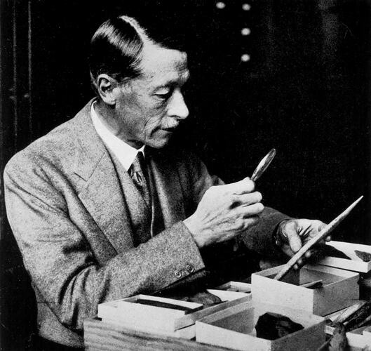 Man wearing a suit and tie seated at a desk inspecting rock specimens with magnifying glass.
