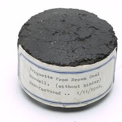 Brown disc-shaped briquette with paper label affixed around side.
