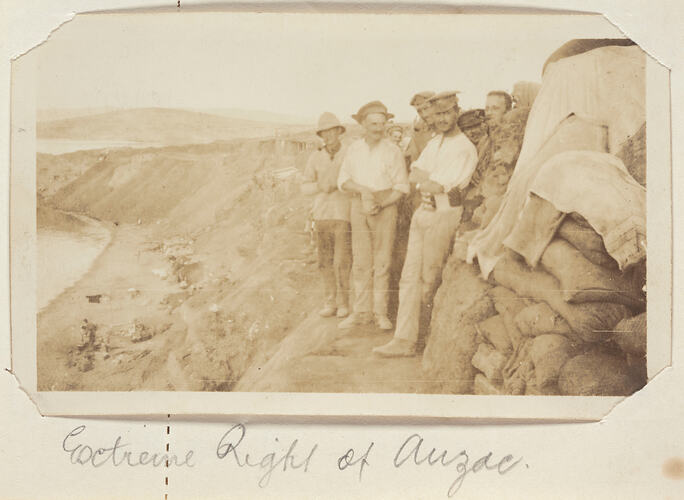 Group of men standing on a ledge of a cliff next to a pile of sand bags, on left a body of water.