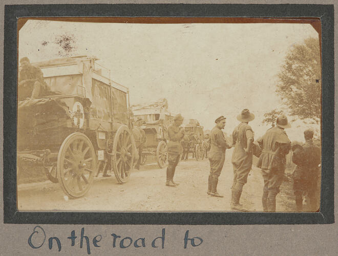 Convoy of horse drawn wagons on dirt road, with group of soldiers standing on the side of the road.
