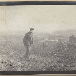 Man with bandaged face walking in left foreground, with huts and horse carts in background.