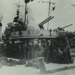 Group of uniformed soldiers standing and sitting in front of a large tank, ships mast in the background.