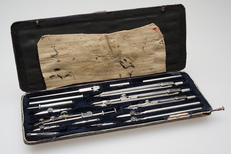 Open box with multiple metal drafting tools. Black lining. Label inside top lid.