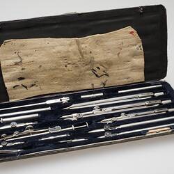 Open box with multiple metal drafting tools. Black lining. Label inside top lid.