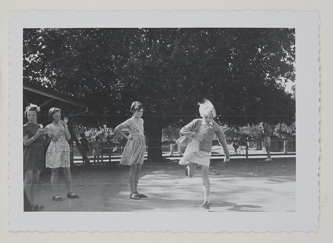 Four schoolgirls playing hopscotch in a playground with a large tree behind them.