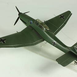 Model aeroplane. Dark green with black cross on side, wing tips and tail. Black propeller. Back view.