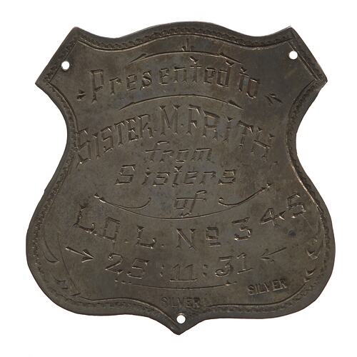 Shield shaped badge with decorative inscription. Has three holes.