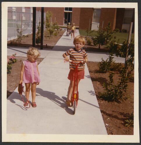 Girl and boy on a footpath outside a building. The boy rides a scooter. He wears red, the girl wears pink.