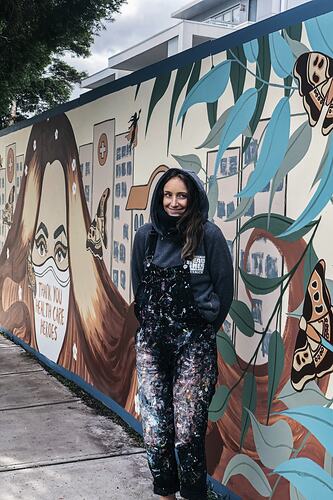 Artist in paint splatered overalls stands in front of colourfully painted wall mural.